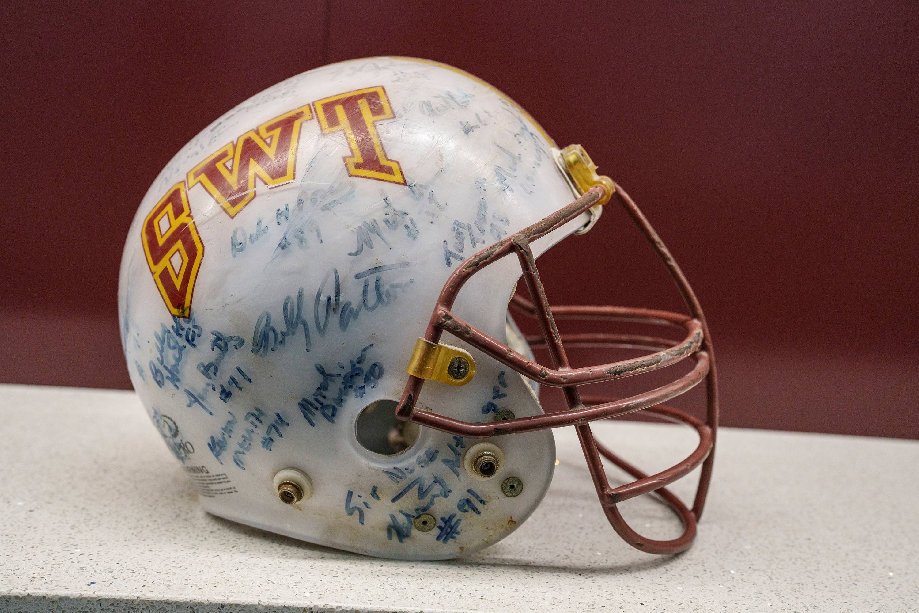 A battered white football helmet with a maroon and gold SWT on the side. It's covered in signatures from the team, signed in faded blue sharpie and sits on a white desk.