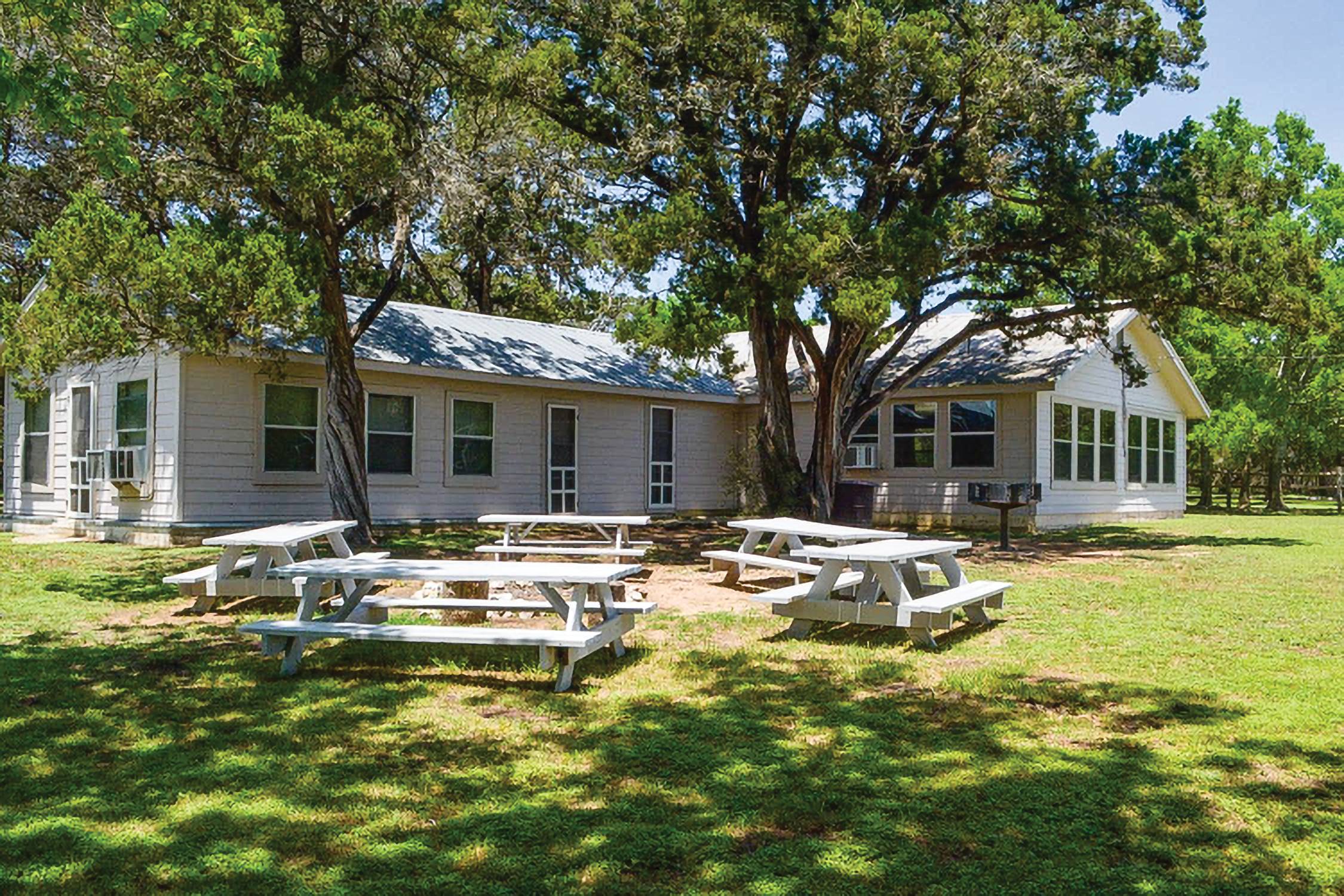 exterior shot of white home with white picnic tables in front of it. there are lots of green lush trees around the home. 