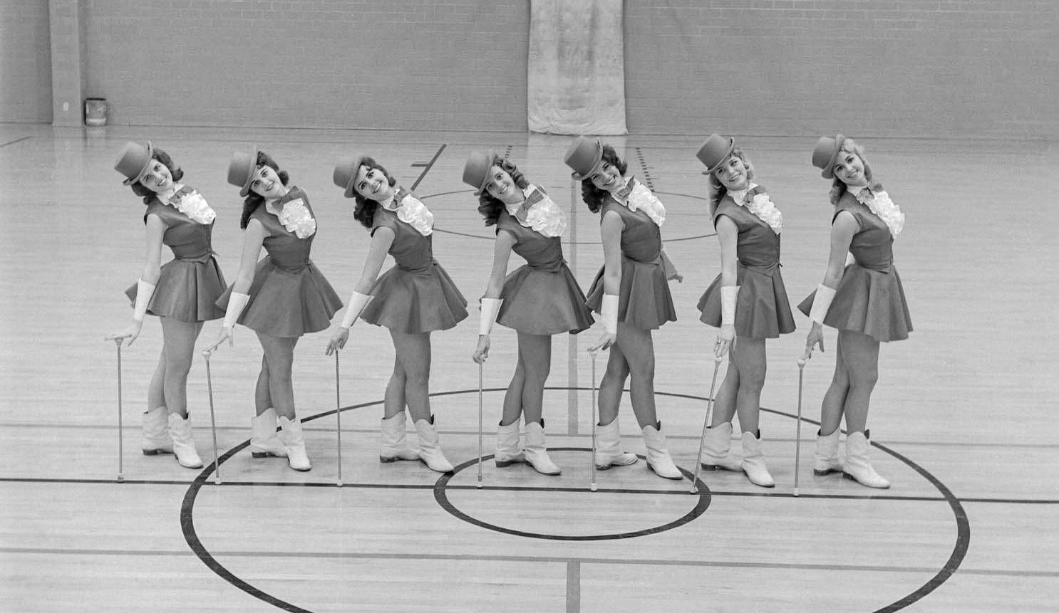 Vintage photo of 7 women posing in matching uniforms of a short skirt, sleeveless tuxedo shirt, and bow tie with short top hats and canes.