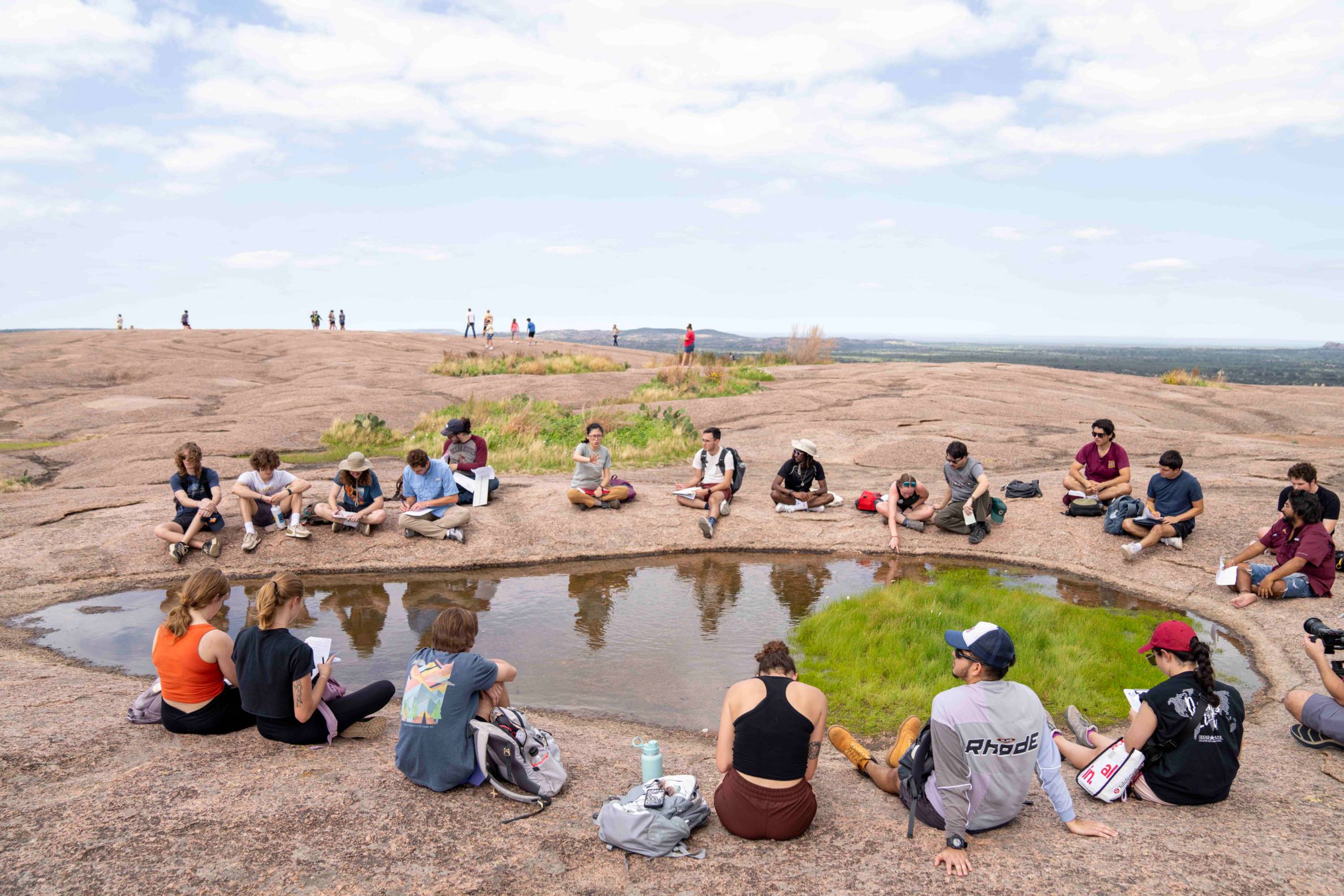 A group of young folks in hiking gear sit cross-legged next to a puddle on a rocky surface.