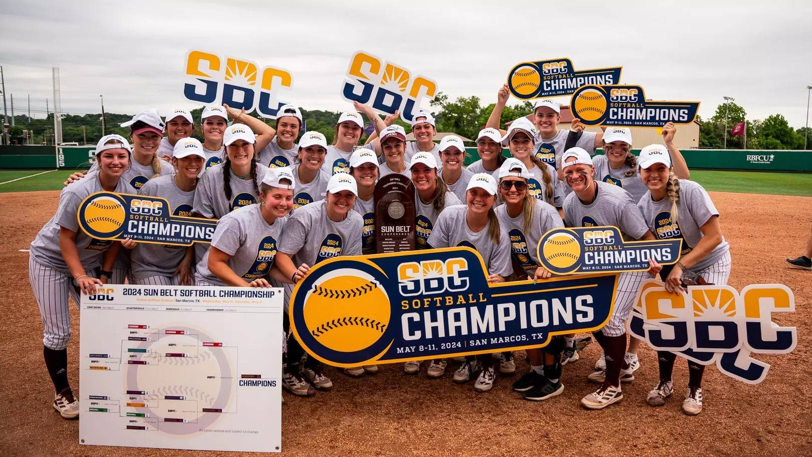 The women's softball team smiles and poses, carrying banners that hold variations of the phrase "SBC Champions".