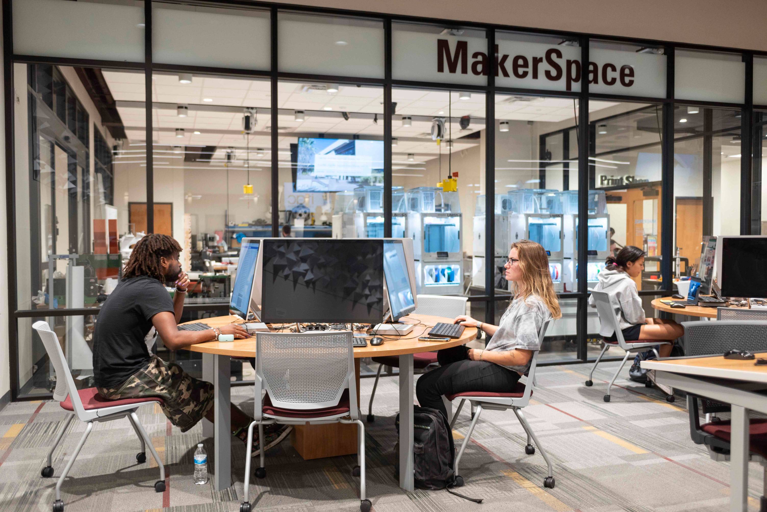 Students sit working at computers. In the background is a wall sign that reads "Maker Space" and a glass room showcasing different equipment.