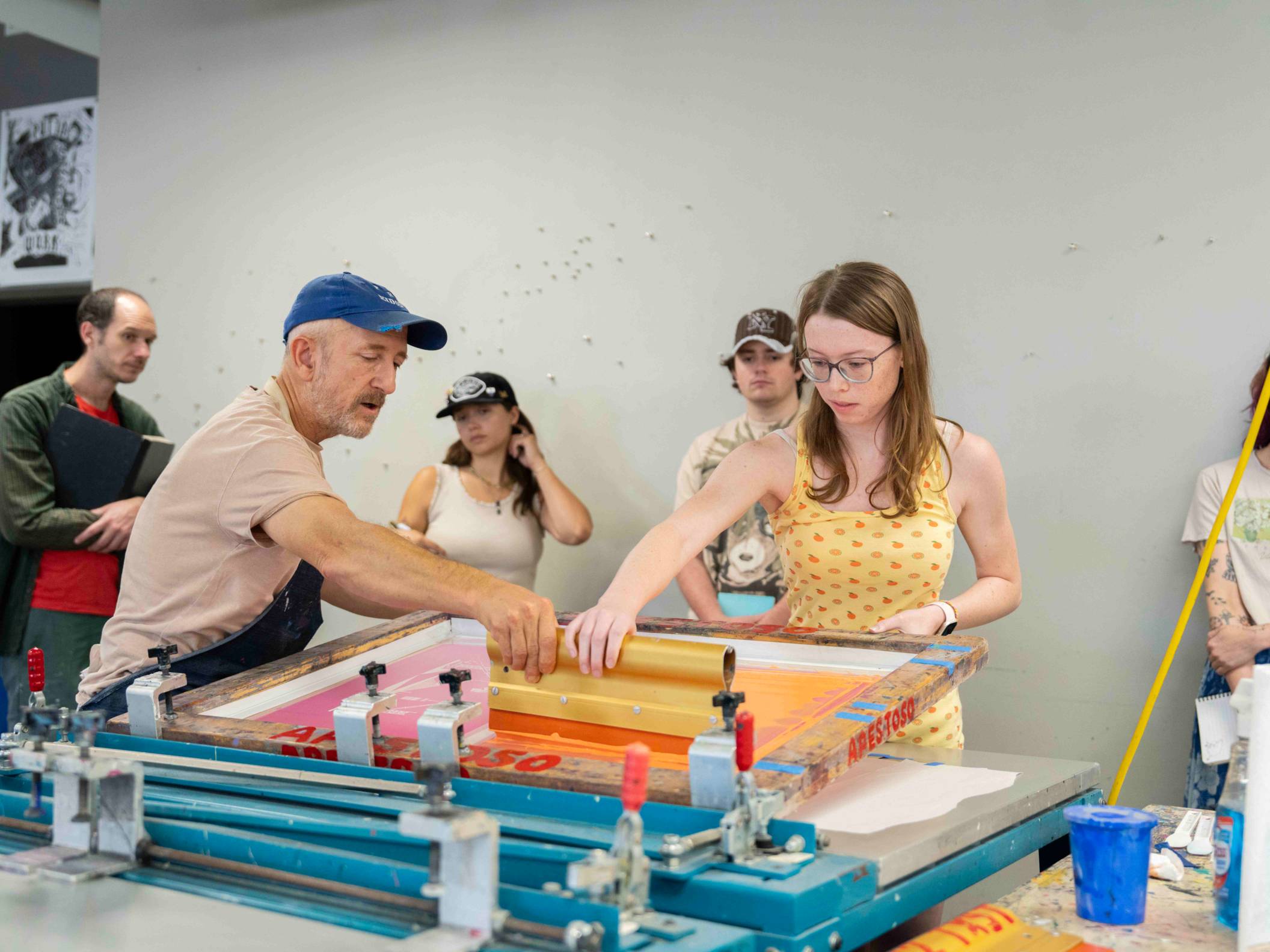 A teacher wearing a hat and apron assists a student as she scrapes paint down a large sheet of paper.