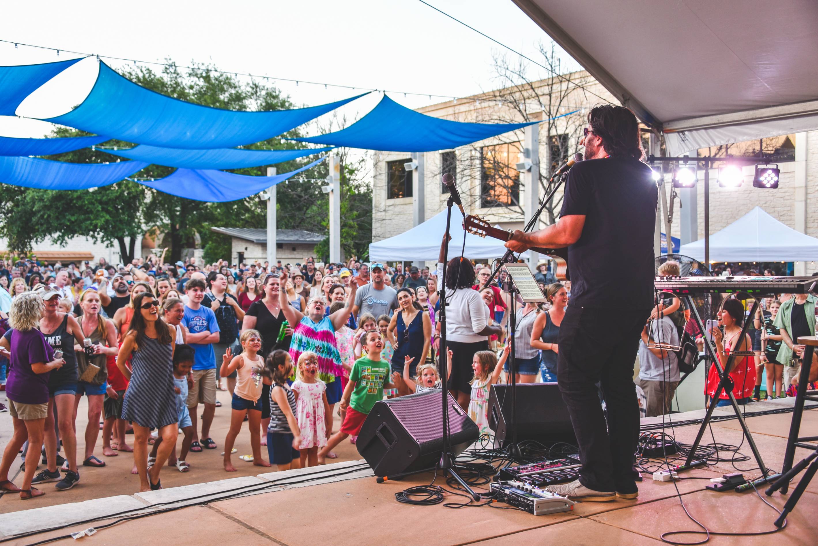 Shot taken from the back of the stage. On stage, a man wearing all black sings and plays guitar. Ahead of him are a range of young folks dancing in the crowd.