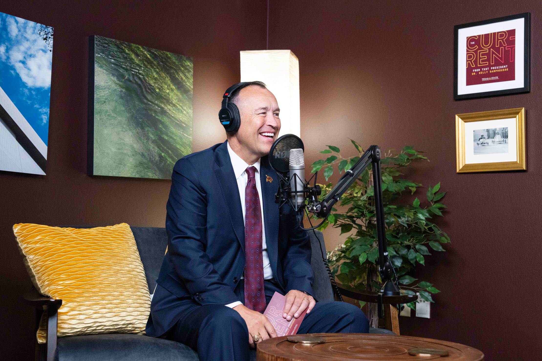 President Damphousse sitting on a yellow couch in a swanky room. He is wearing headphones and laughing into a microphone.