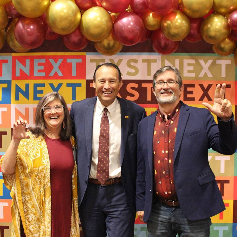 Dee and Doug Bynum smile for a celebratory photograph in front of a colorful backdrop at an event with Kelly Damphousse.