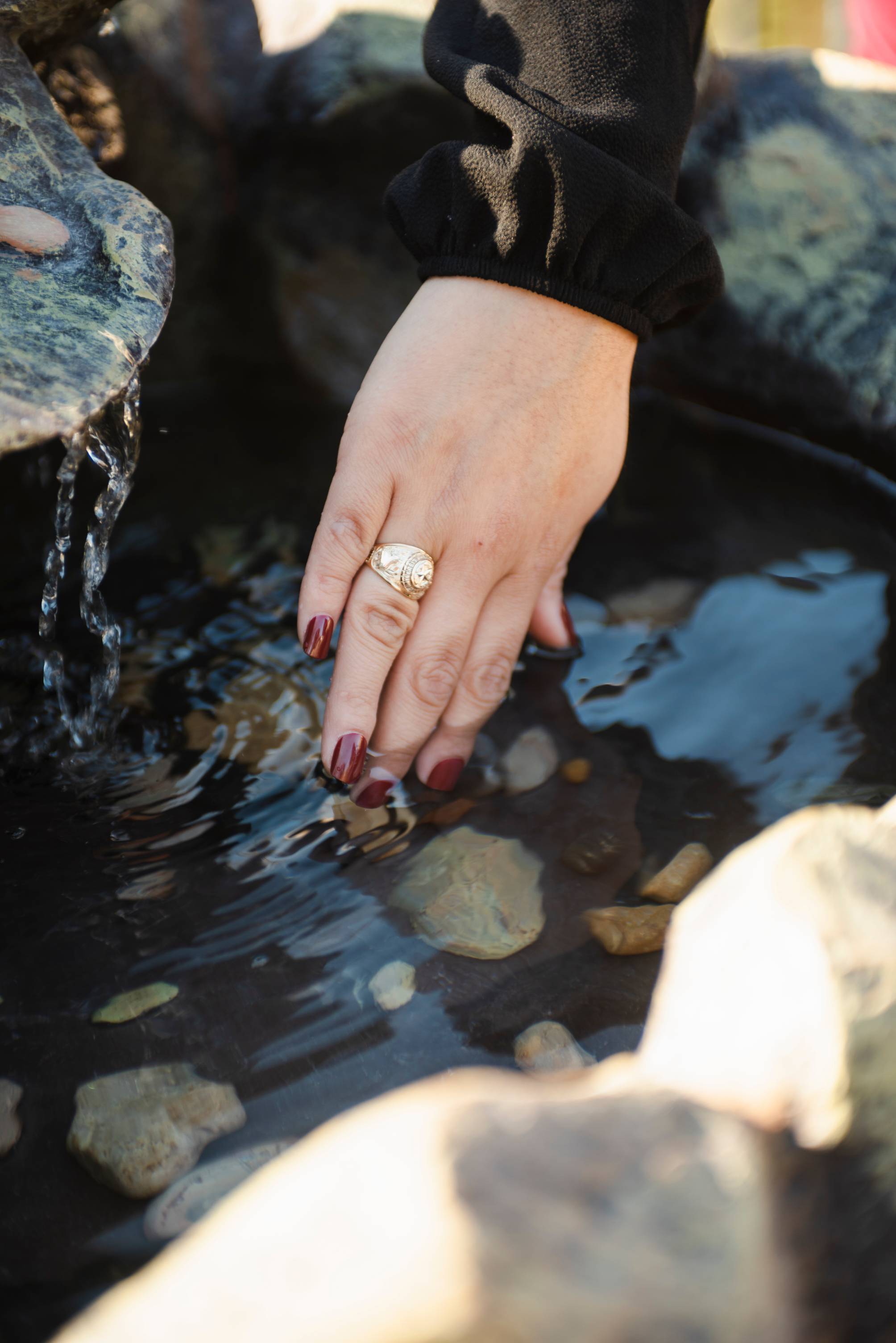 A woman's hand wearing a gold class ring dips fingers into a small pool of water.