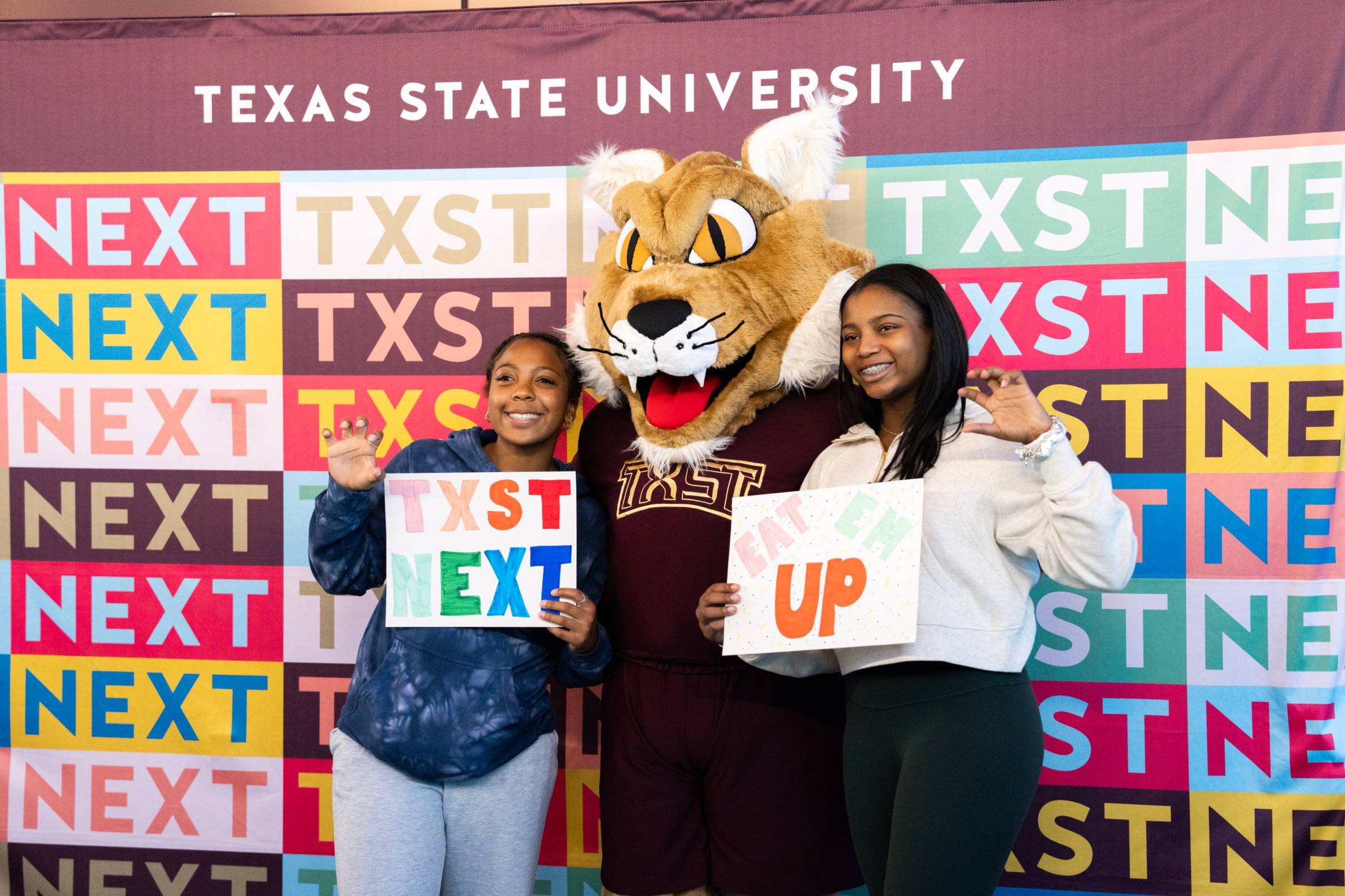 bobcat mascot poses with two women in front of color step-and-repeat that reads TXST NEXT