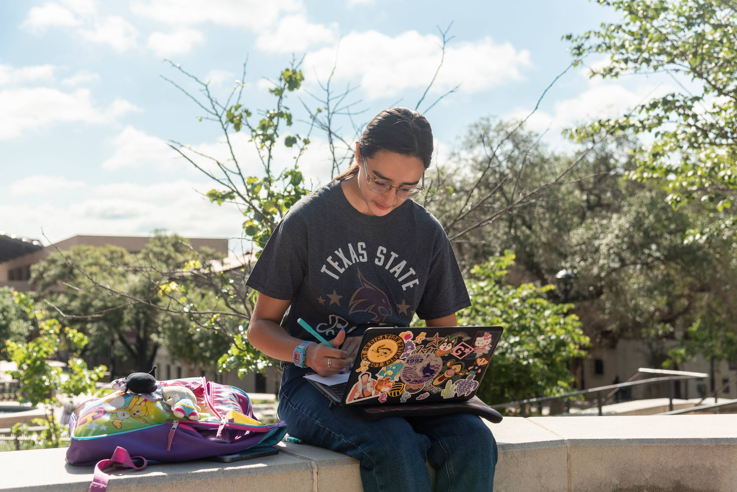 A female student sits on a ledge with her laptop on her lap. The laptop is covered in stickers and she is writing on an index card.