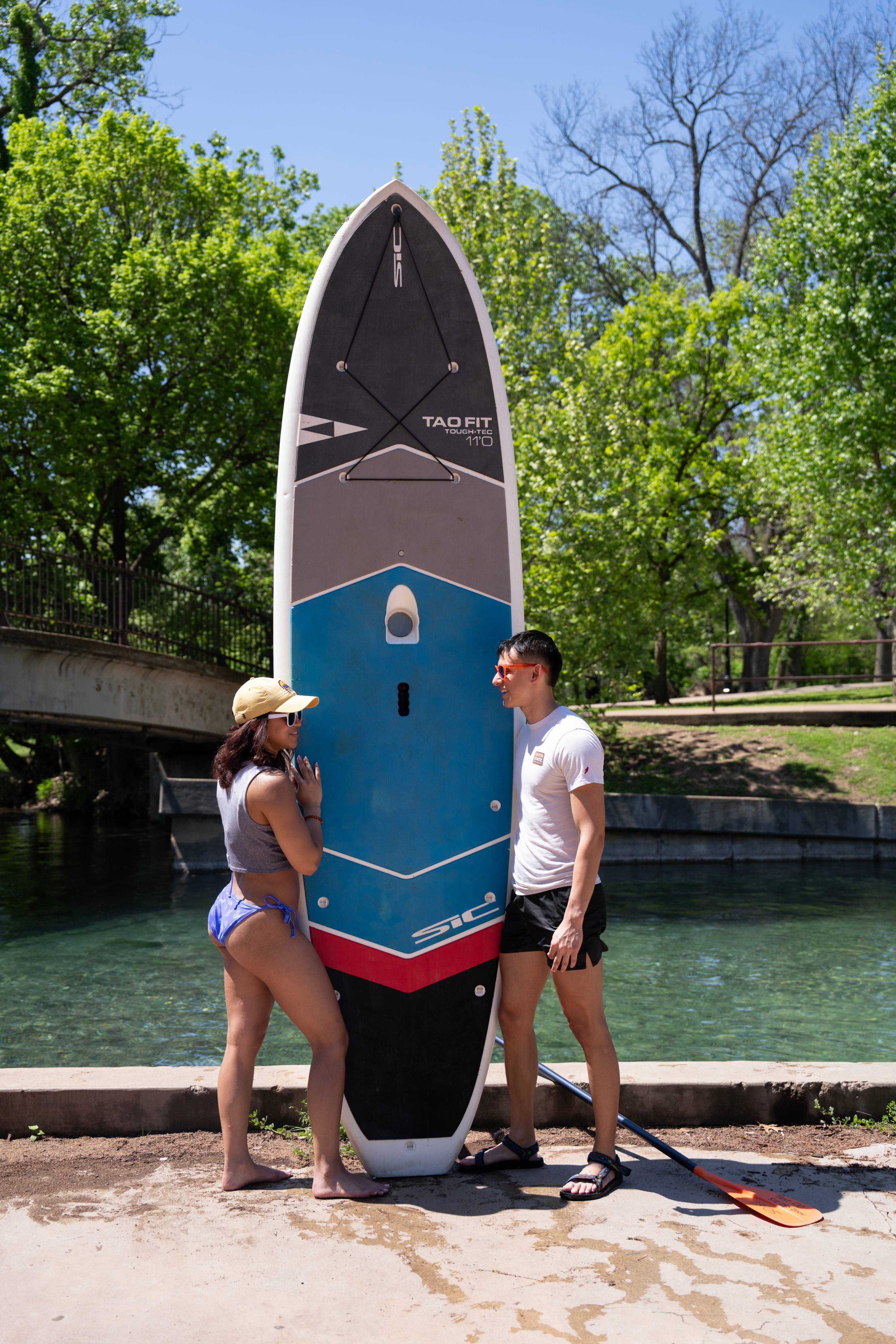 two people pose with a standing paddleboard next to a clear blue river