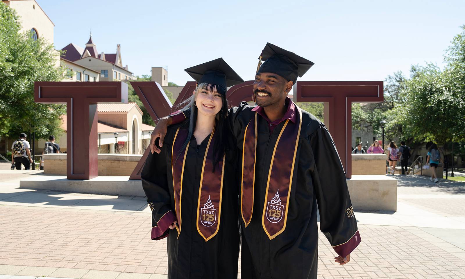 A student poses for a graduation portrait wearing their graduation regalia, including a Texas State 125th anniversary stole.