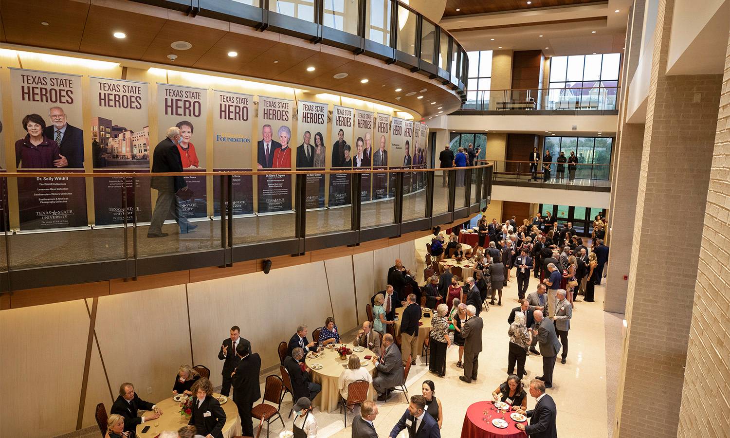 A crowd of well-dressed donors, faculty and staff mingle at the 2021 Capital Campaign Gala event. Banners honor top-level donors, designating them as Texas State Heroes. Old Main can be seen peeking through the window in the background. 