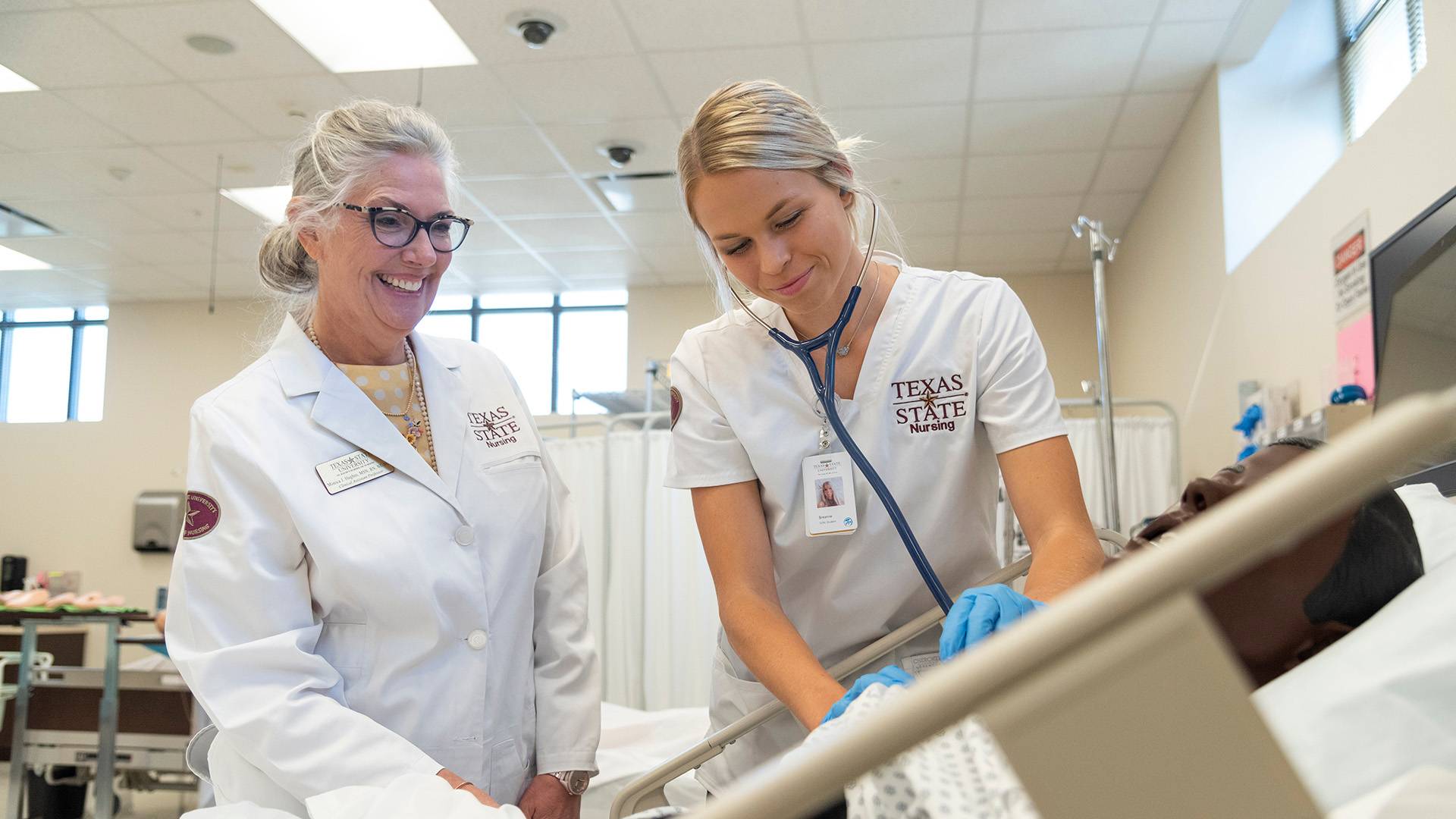 A nursing faculty member looks on while a student performs a test procedure on a dummy during a nursing lab.