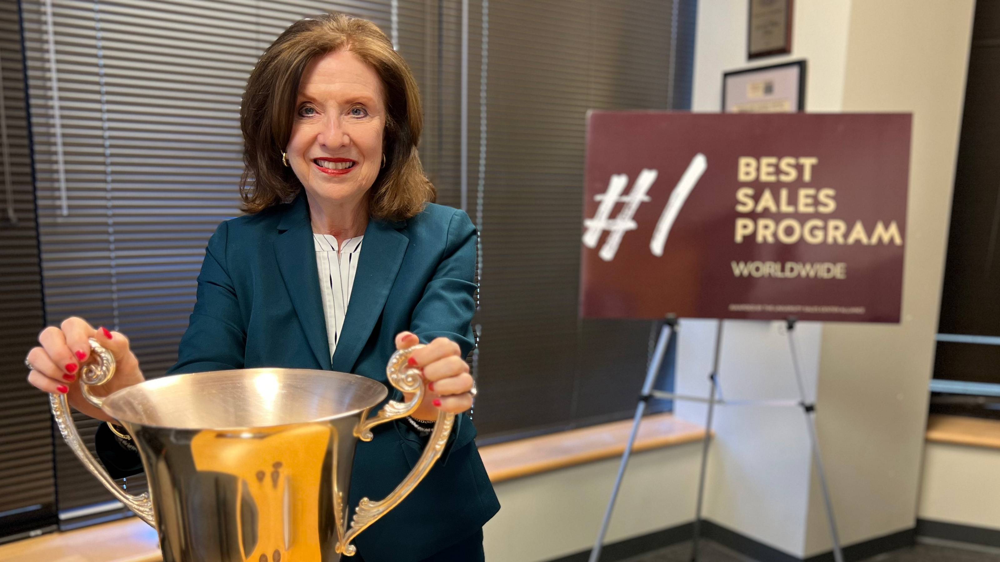 Vicki West in business attire holding onto a large silver cup trophy with sign in background that reads, "Number one sales center worldwide."