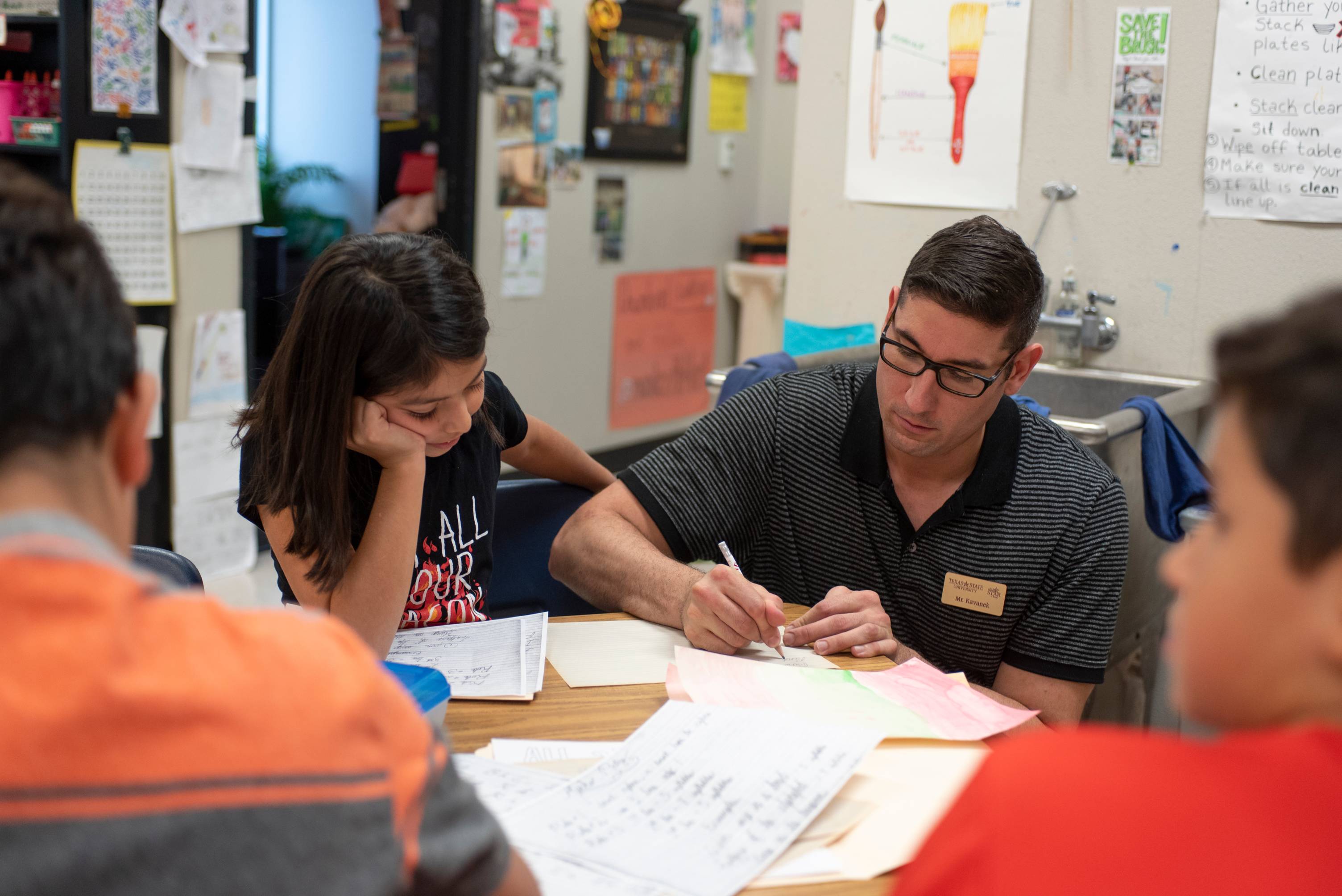 male kneeling next to small table helping young child with her classwork