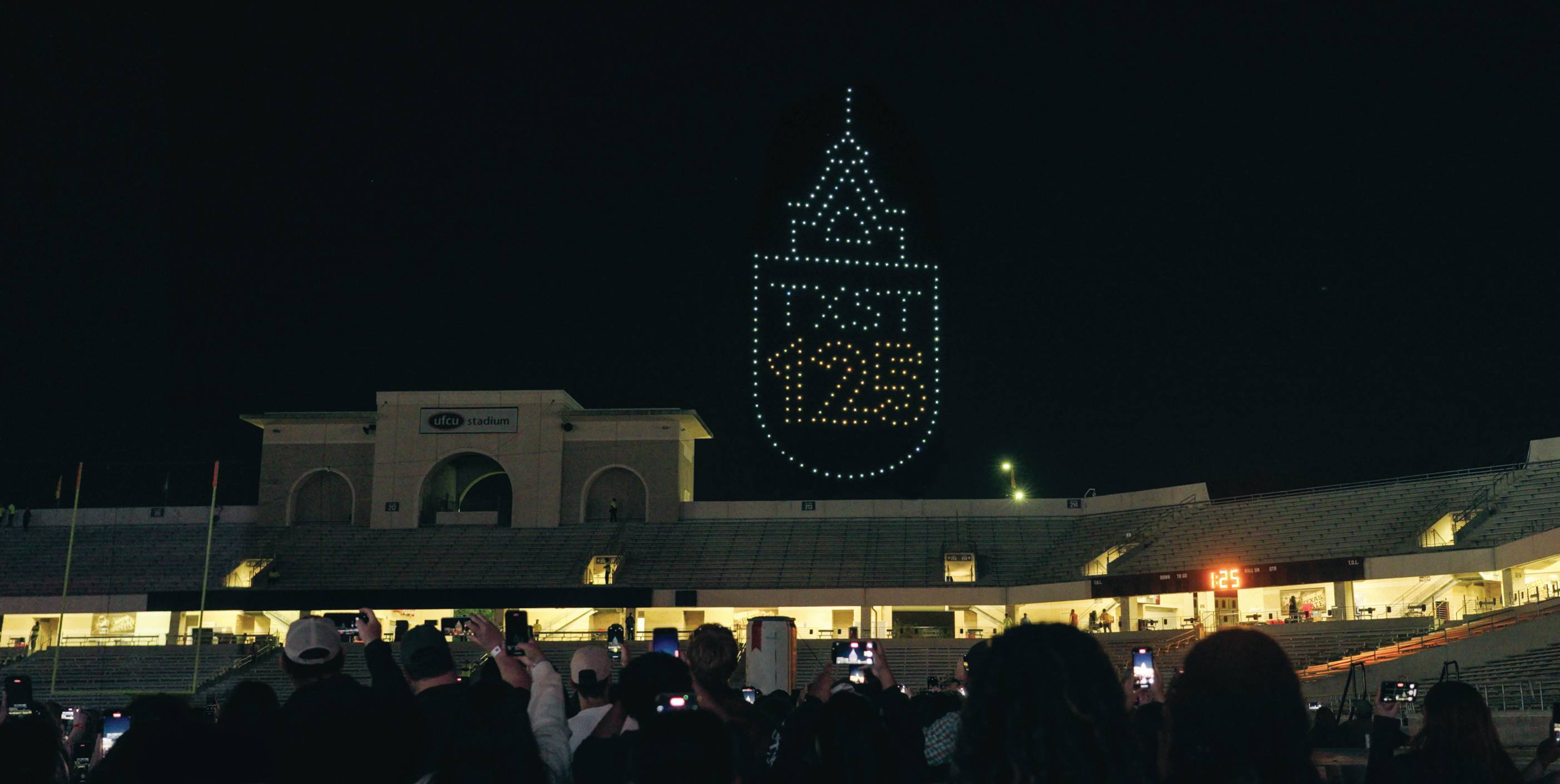 Snapshot of the drone show at UFCU Stadium during the Celebrate 125 concert. A crowd of students, faculty, staff and community members watch and take photos as the drones form the words 'TXST 125.'