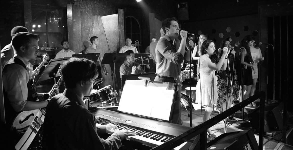 Black and white photo of the jazz ensemble, Salsa del Rio, performing in 1995. Shown are vocalists, a keyboard player, rhythm guitarists, and a brass section.