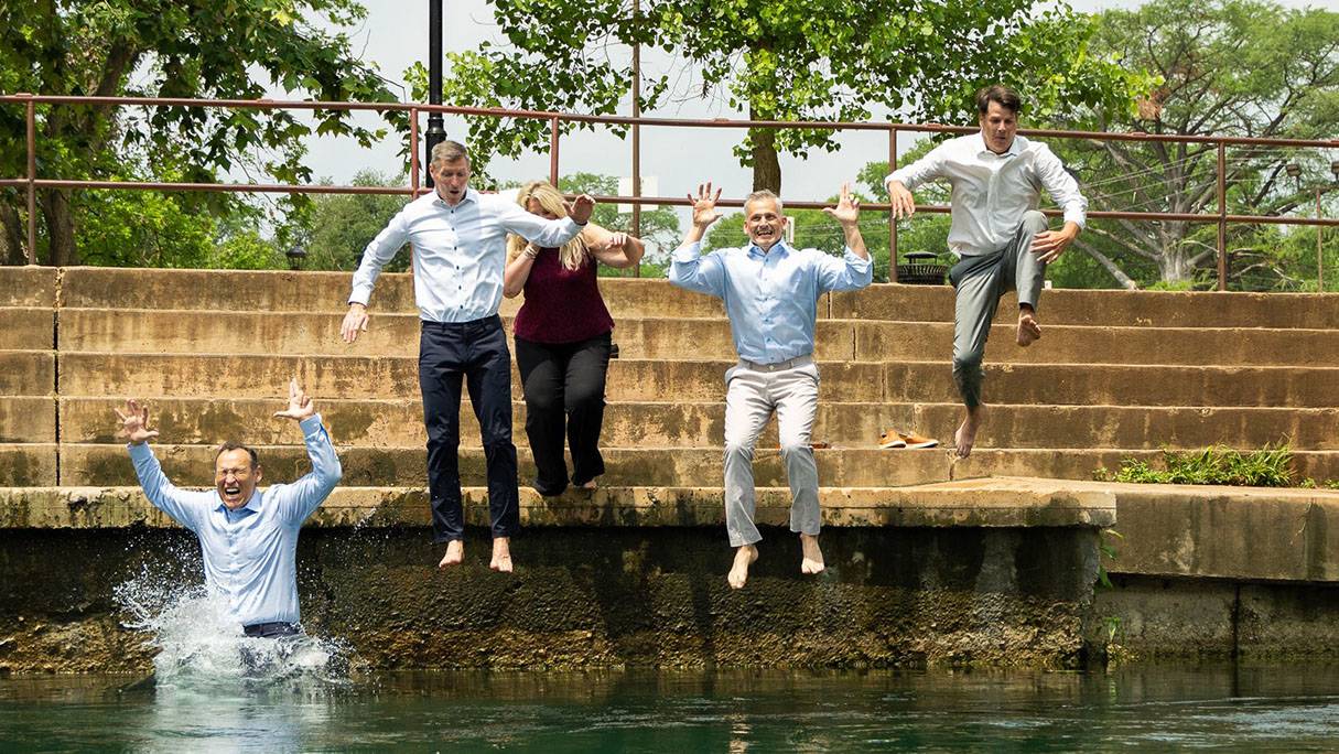 A group of TXST Athletics staff members and UFCU employees jump in the river with President Kelly Damphousse (left).