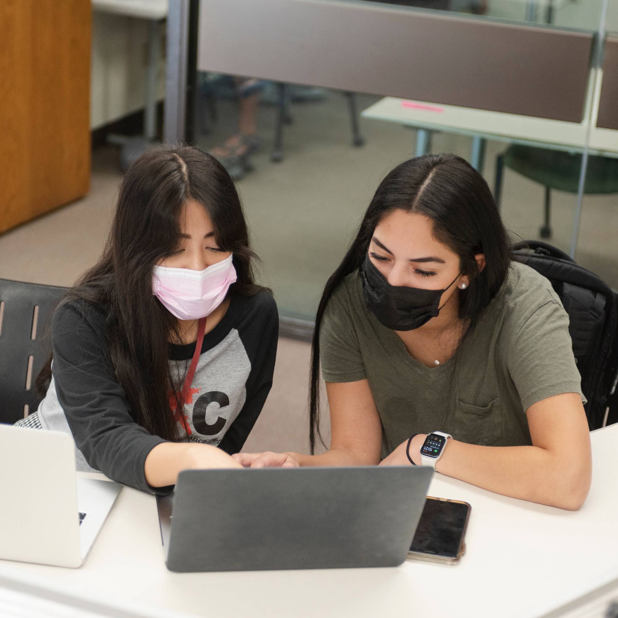 Two students wearing medical masks sit at a table at the library, looking at a laptop. 