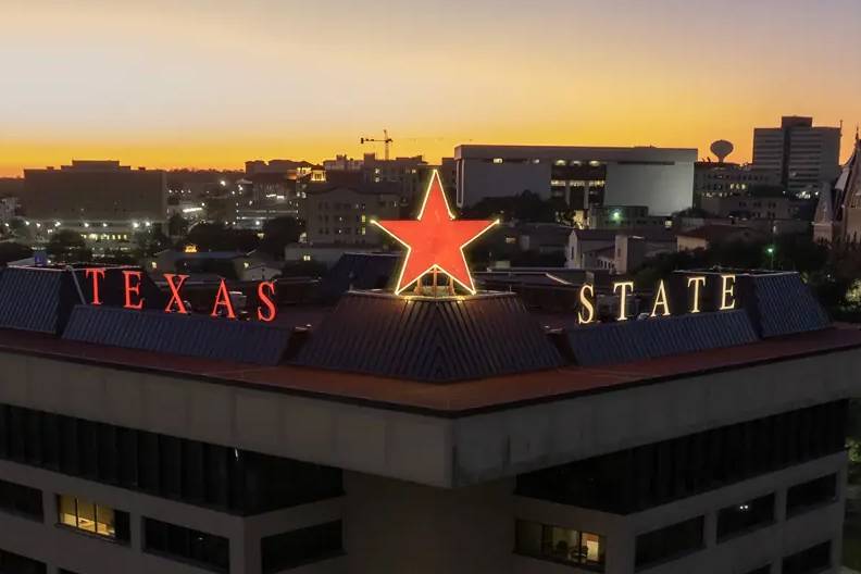 The Victory Star glows on top of the JC Kellam building.  Both the star and "Texas State" are lit up in maroon. 