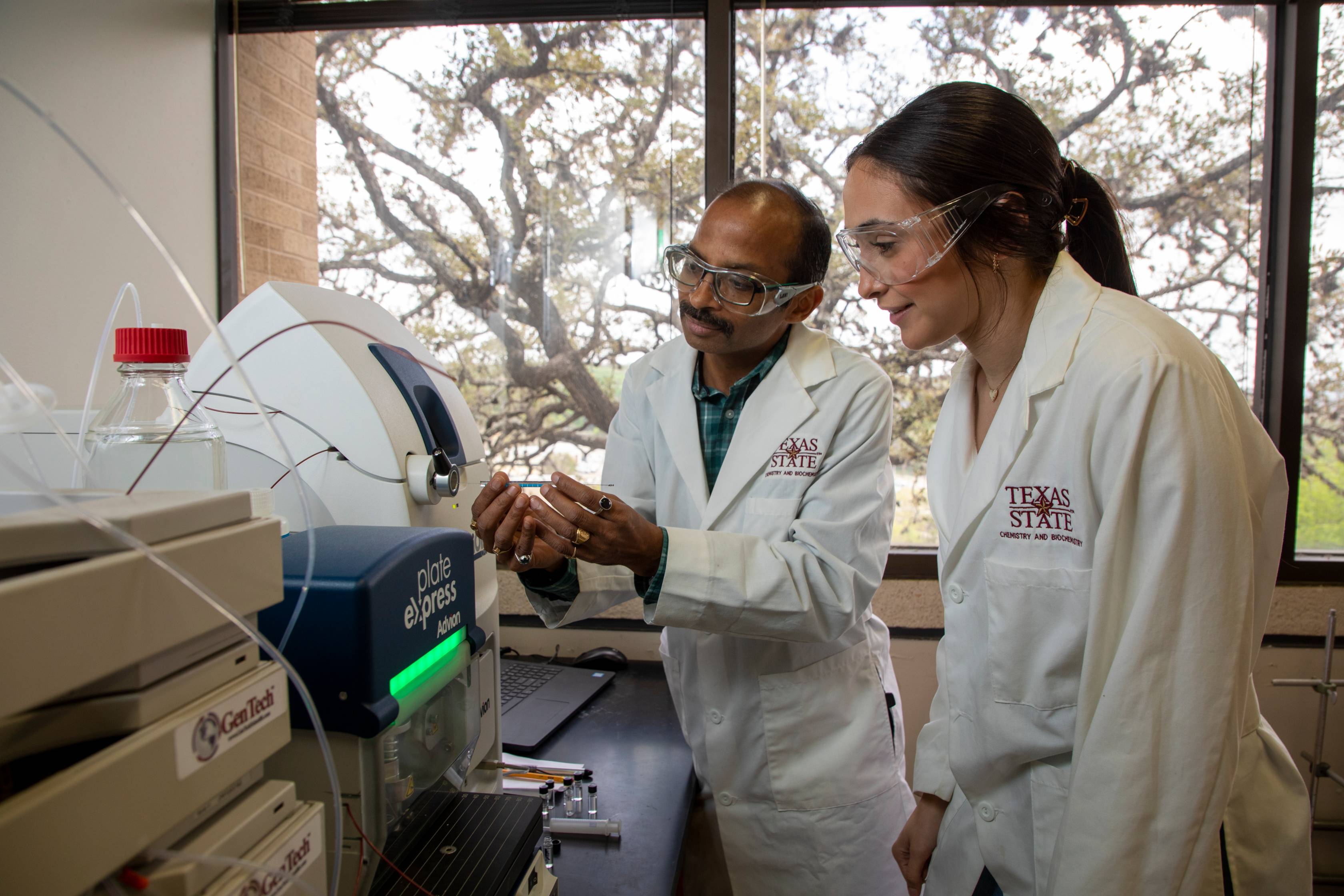 Two researchers in Texas State University lab coats and safety goggles work together in a laboratory. One person is explaining or demonstrating something while holding a small object, and the other listens attentively. 