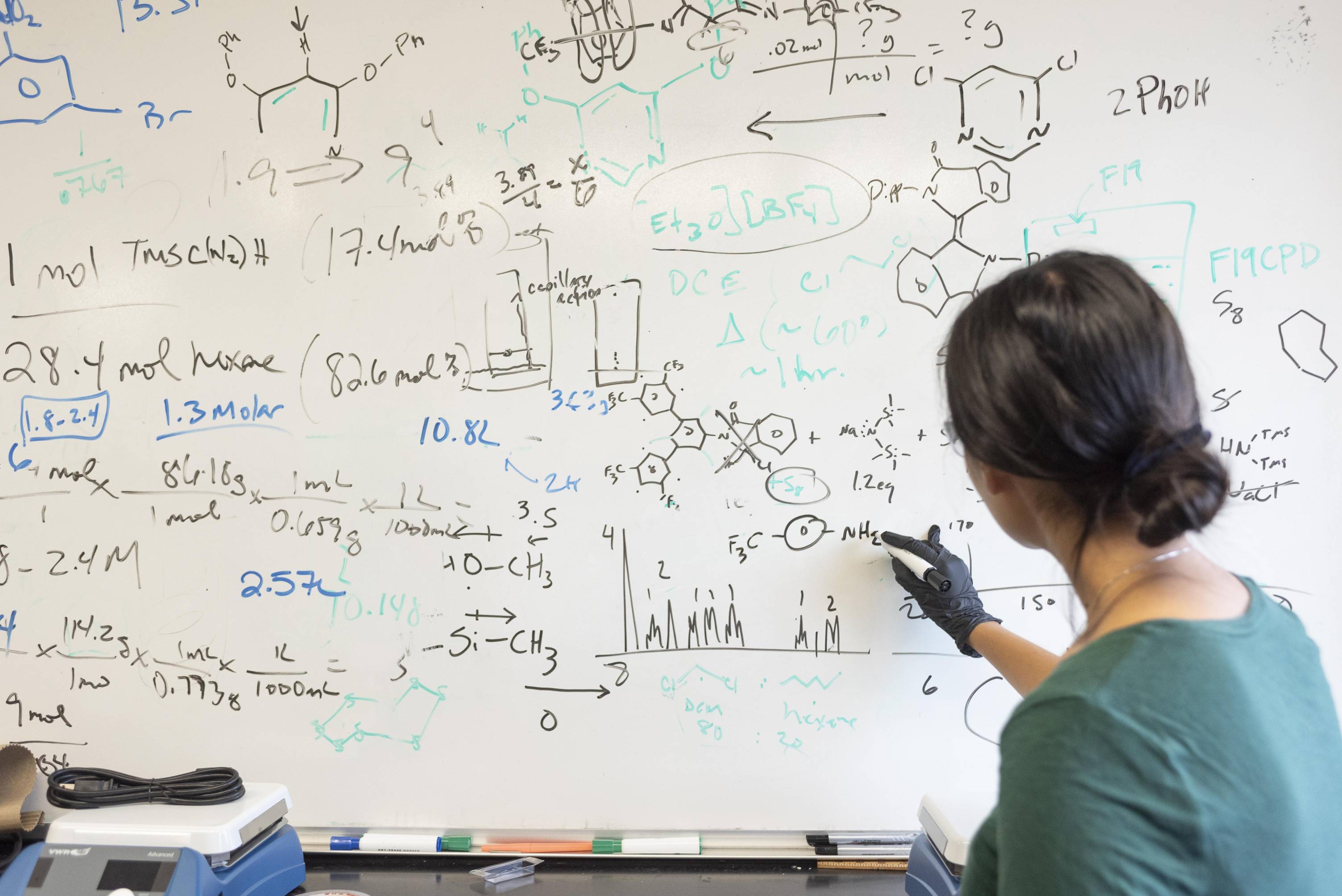 A person with dark hair tied back is writing on a large whiteboard covered with complex chemical structures, equations, and scientific notes. They are wearing a green shirt and a black glove on one hand, working intently in a laboratory or academic setting.