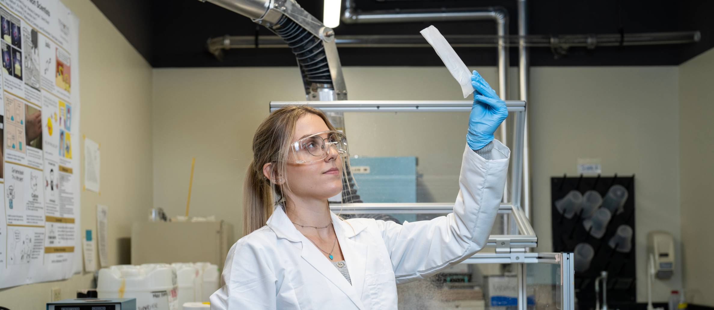 woman in white lab coat and safety glasses holds up a scientific instrument to examine it