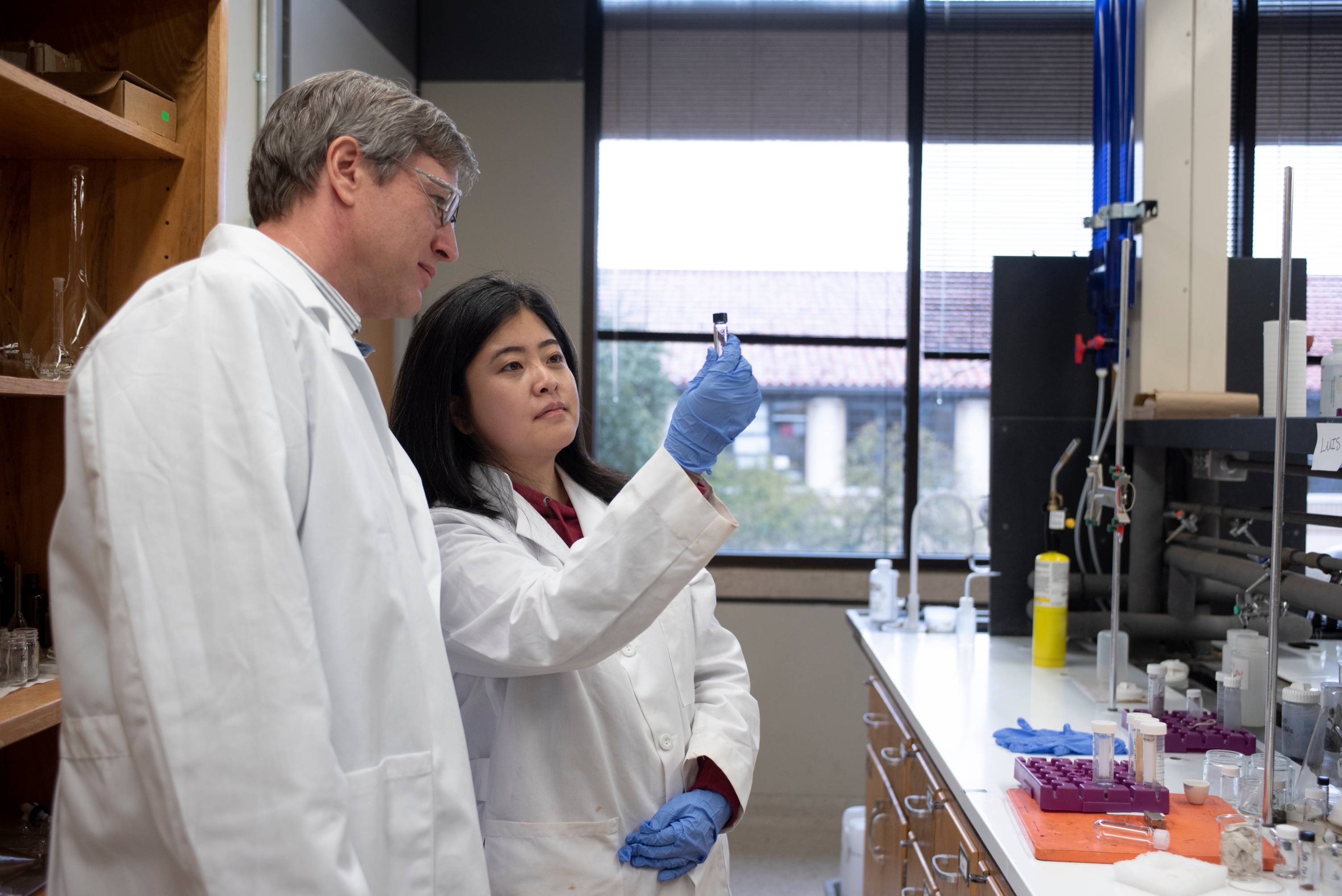 A student in a white lab coat and blue gloves holds up a small vial with dark contents, examining it, while a professor in a white lab coat and glasses looks on.