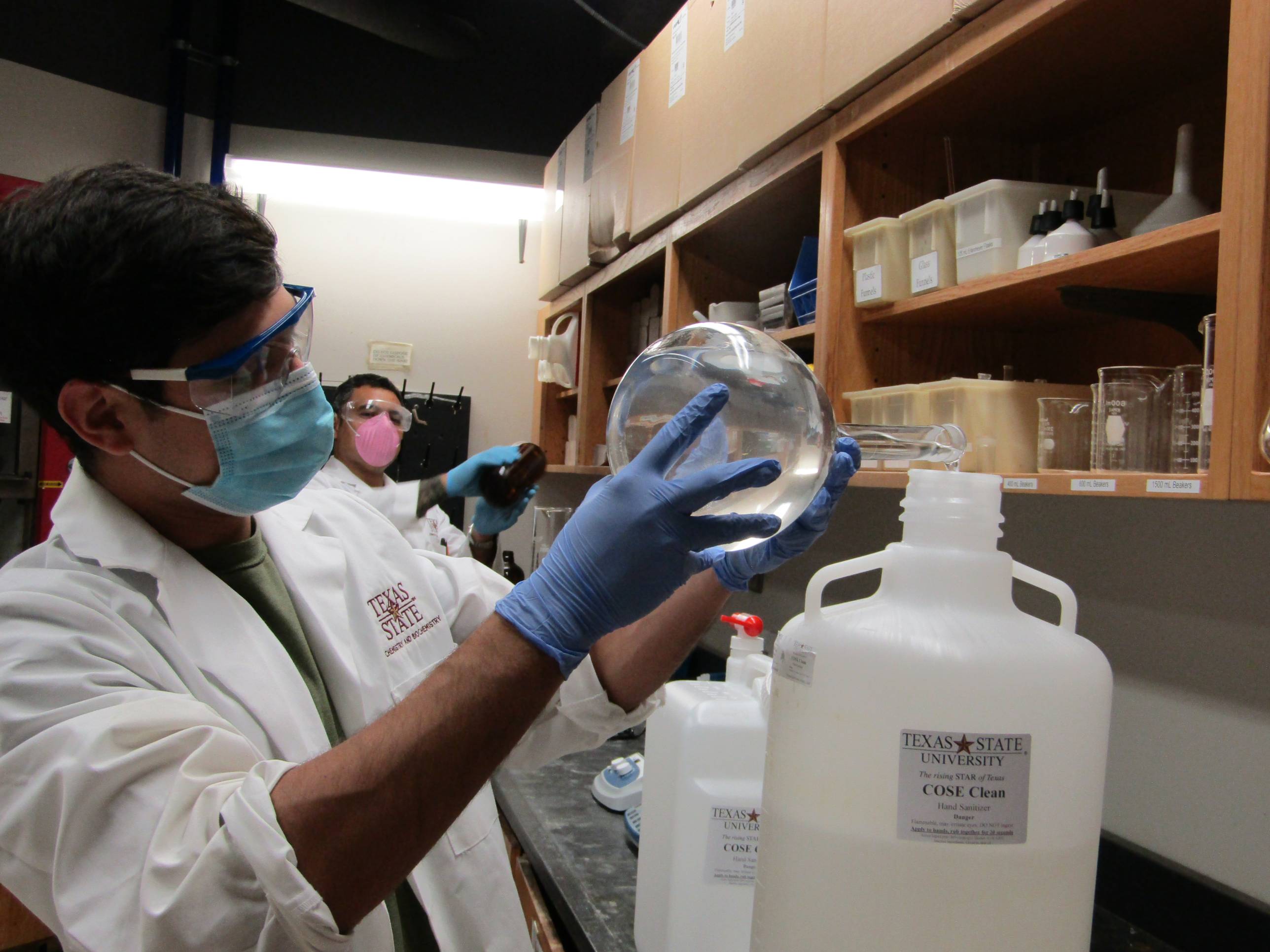 A person wearing a lab coat, blue gloves, a face mask, and safety goggles holds a large, clear glass flask. Another person in similar lab attire is blurred in the background.