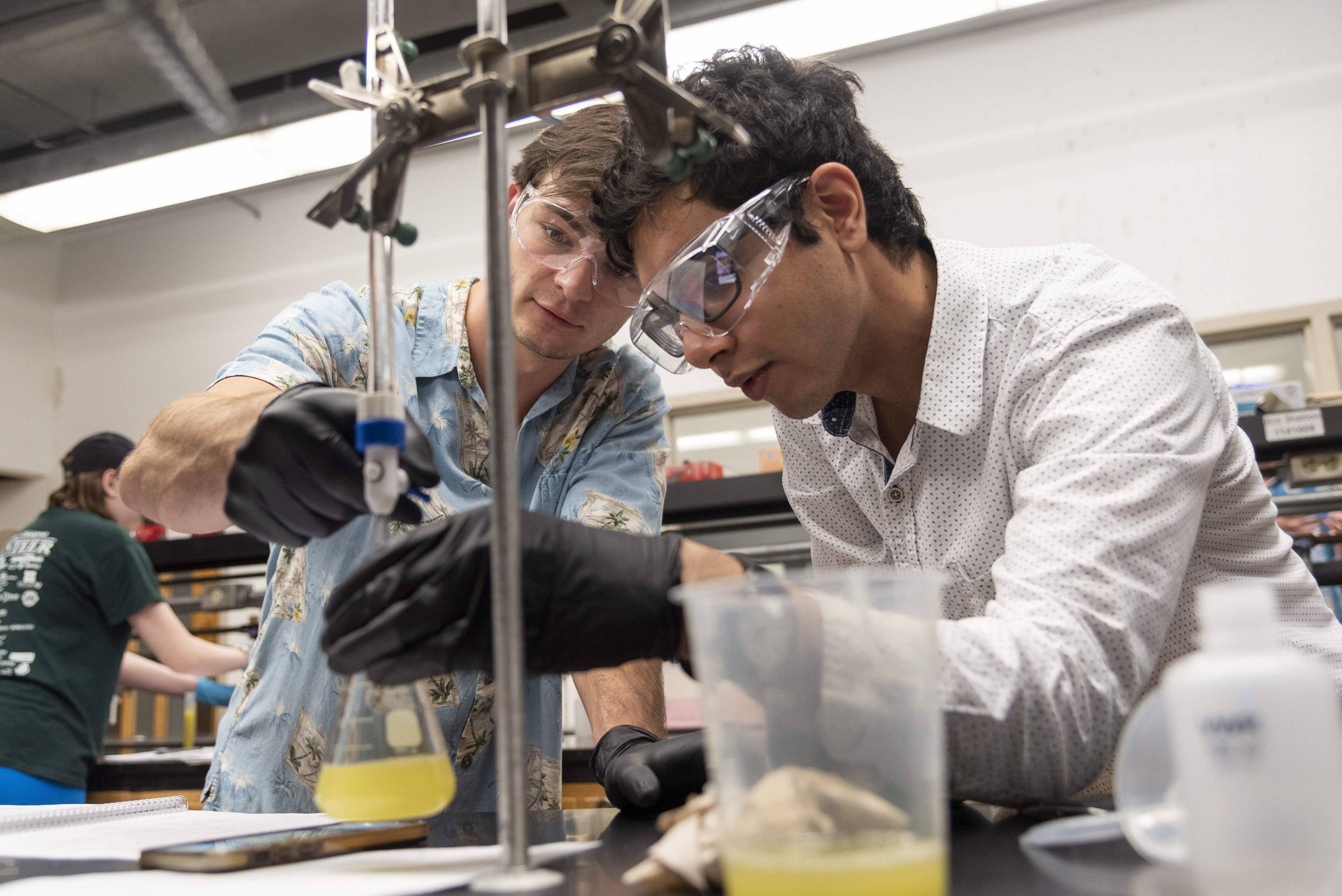 Two students wearing safety goggles and black gloves work together in a laboratory. One holds a glass Erlenmeyer flask containing a yellow liquid while the other adjusts a piece of lab equipment attached to a vertical stand. 