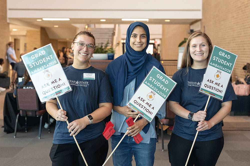 Three graduate student ambassadors smiling while holding up signs signaling that they can be asked questions