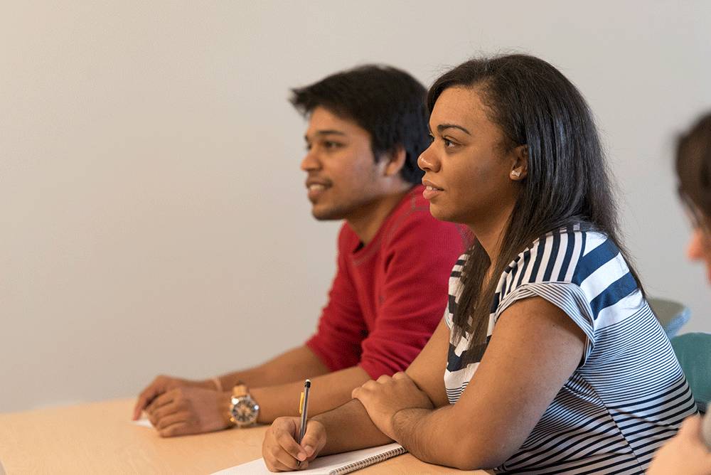 two students sit at a table smiling as they listen to a presentation