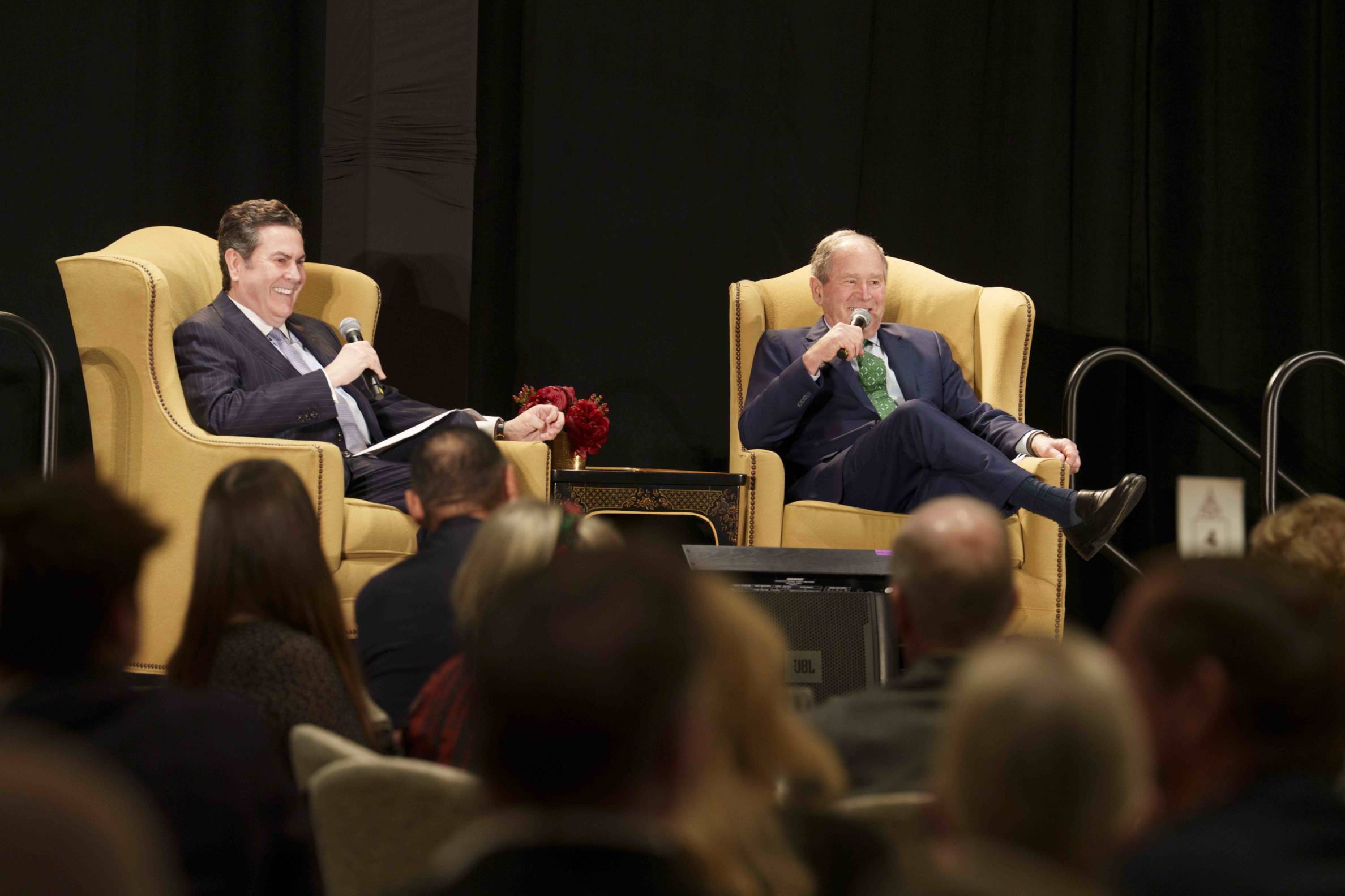 Two men sit on stage talking while sitting in yellow armchairs