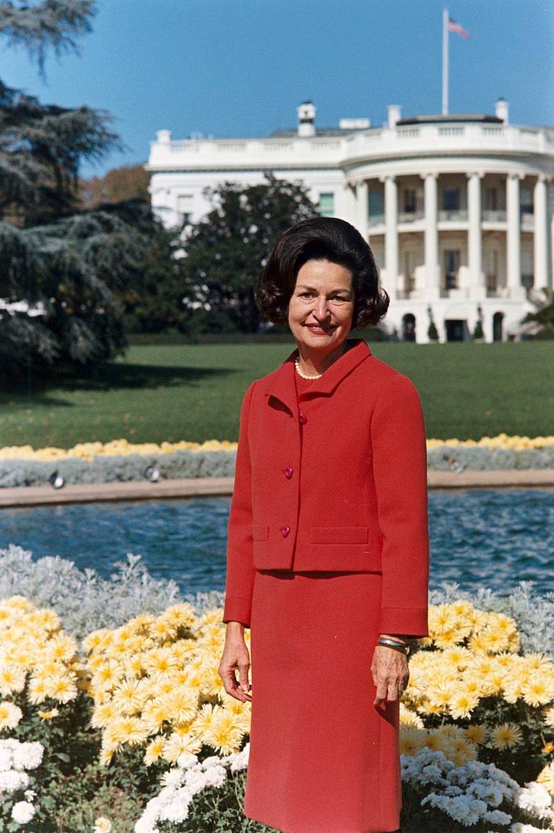 Lady Bird Johnson stands in front of the White House in a vibrant red two-piece