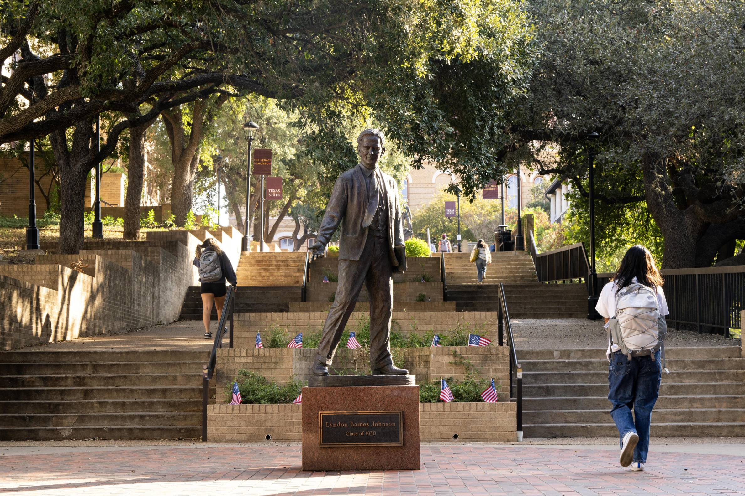 Statue of Lyndon Baines Johnson at Texas State University, with students walking up and down the stairs surrounded by trees and campus banners.