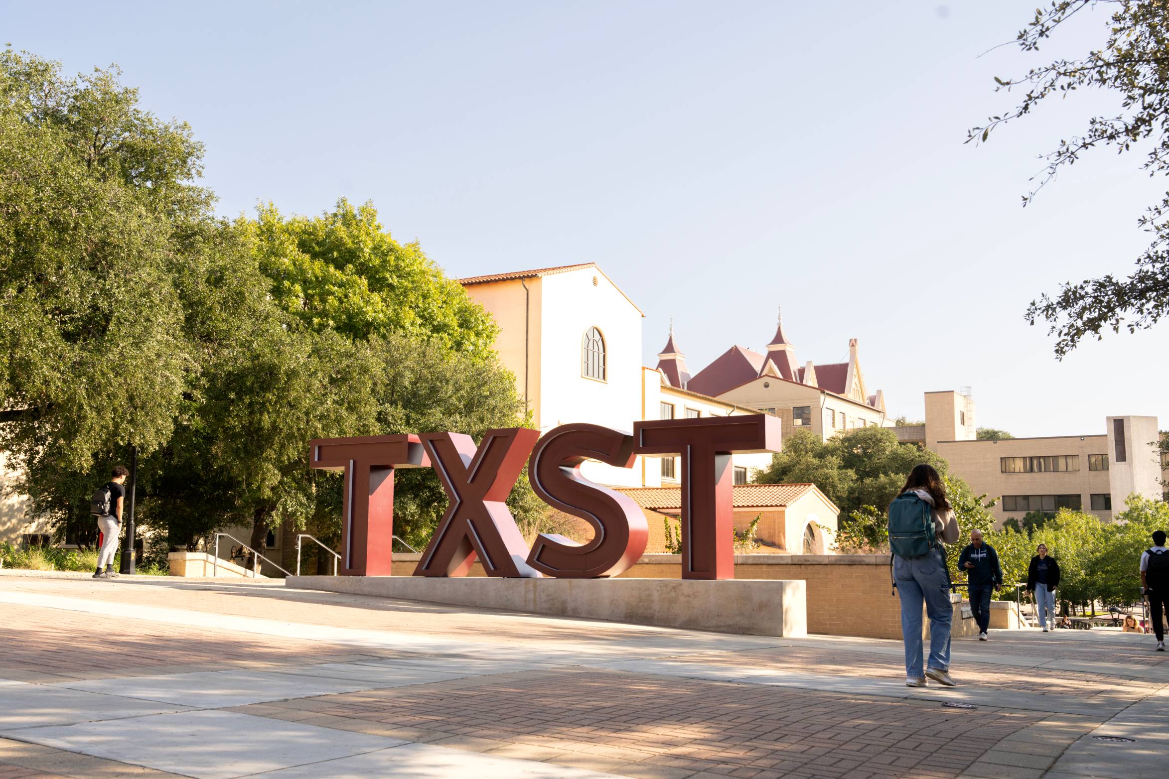 A large, reddish-brown "TXST" sculpture stands prominently on a paved area with students walking around, set against a backdrop of green trees and university buildings under a clear sky.