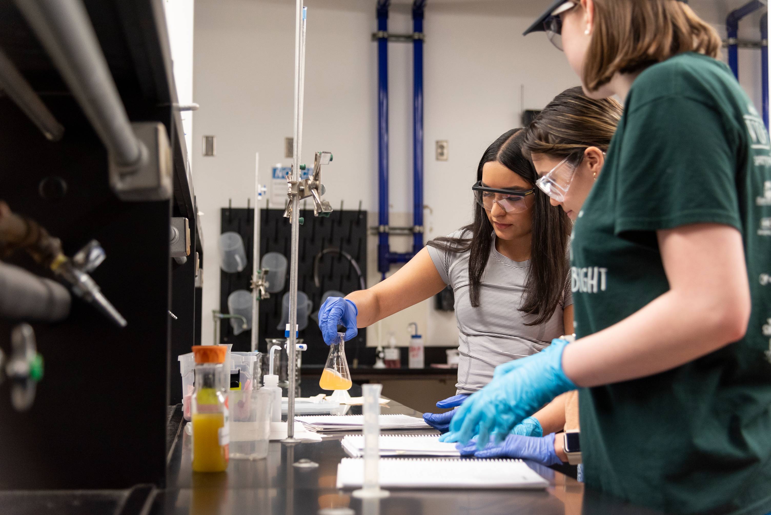  Three students, two wearing safety goggles and blue gloves, are in a chemistry lab. One student holds a flask with a yellow liquid, while the others watch.