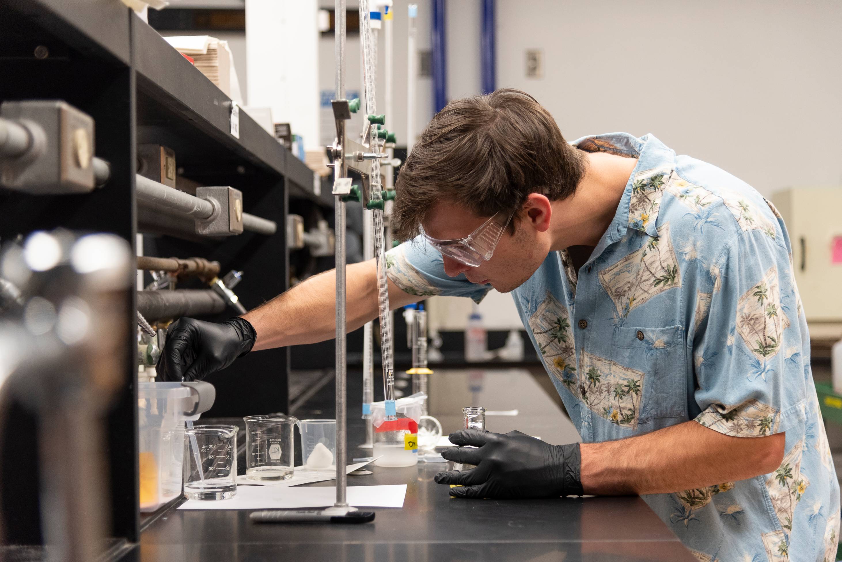 A student in the chemistry lab with goggles and gloves on and a Hawaiian shirt conducting an experiment.