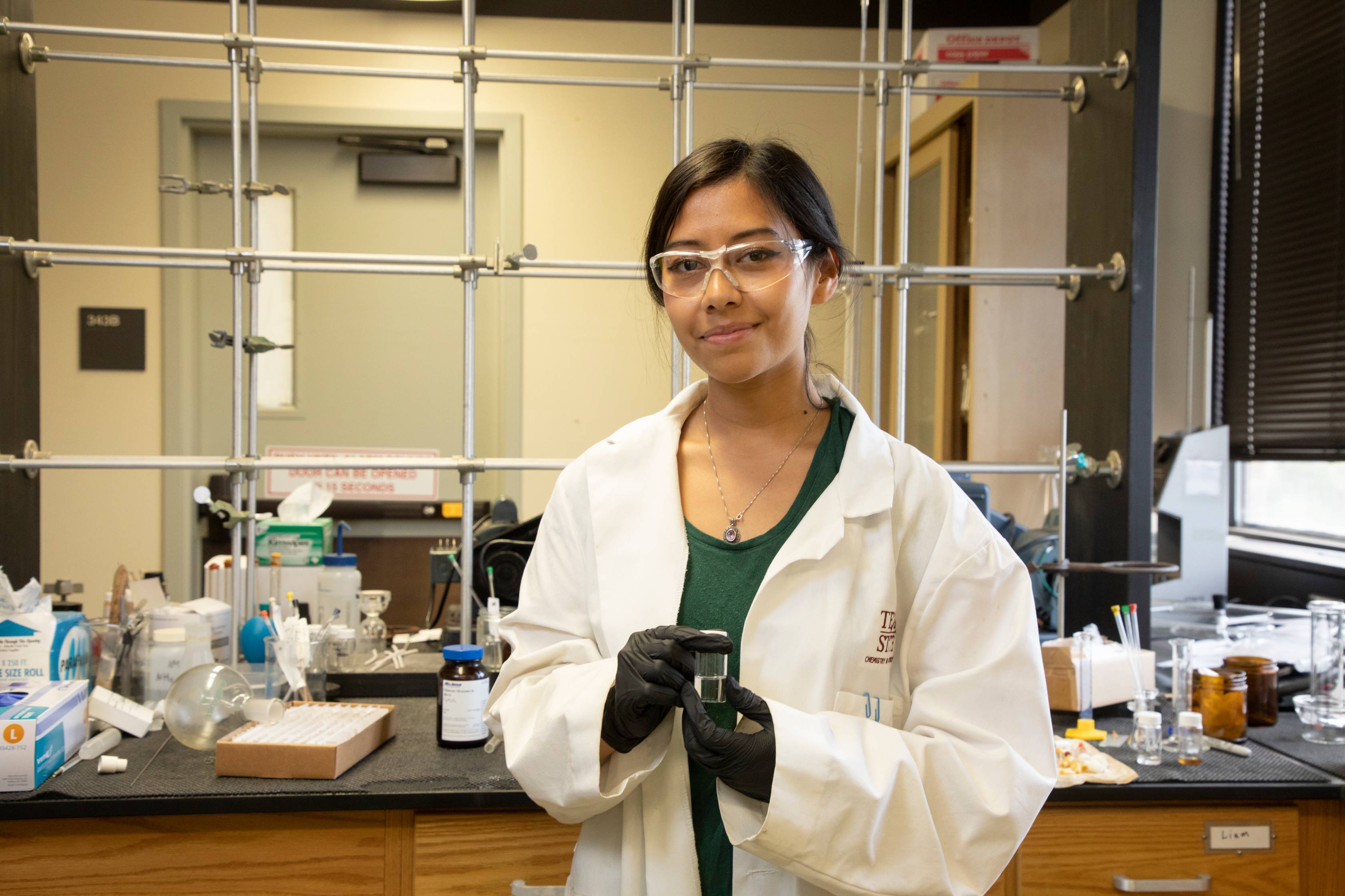 A student wearing a white lab coat, safety glasses, and black gloves smiles at the camera while holding a clear glass vial in a chemistry lab. The lab features shelves with glassware, bottles, and a metal rack structure in the background.