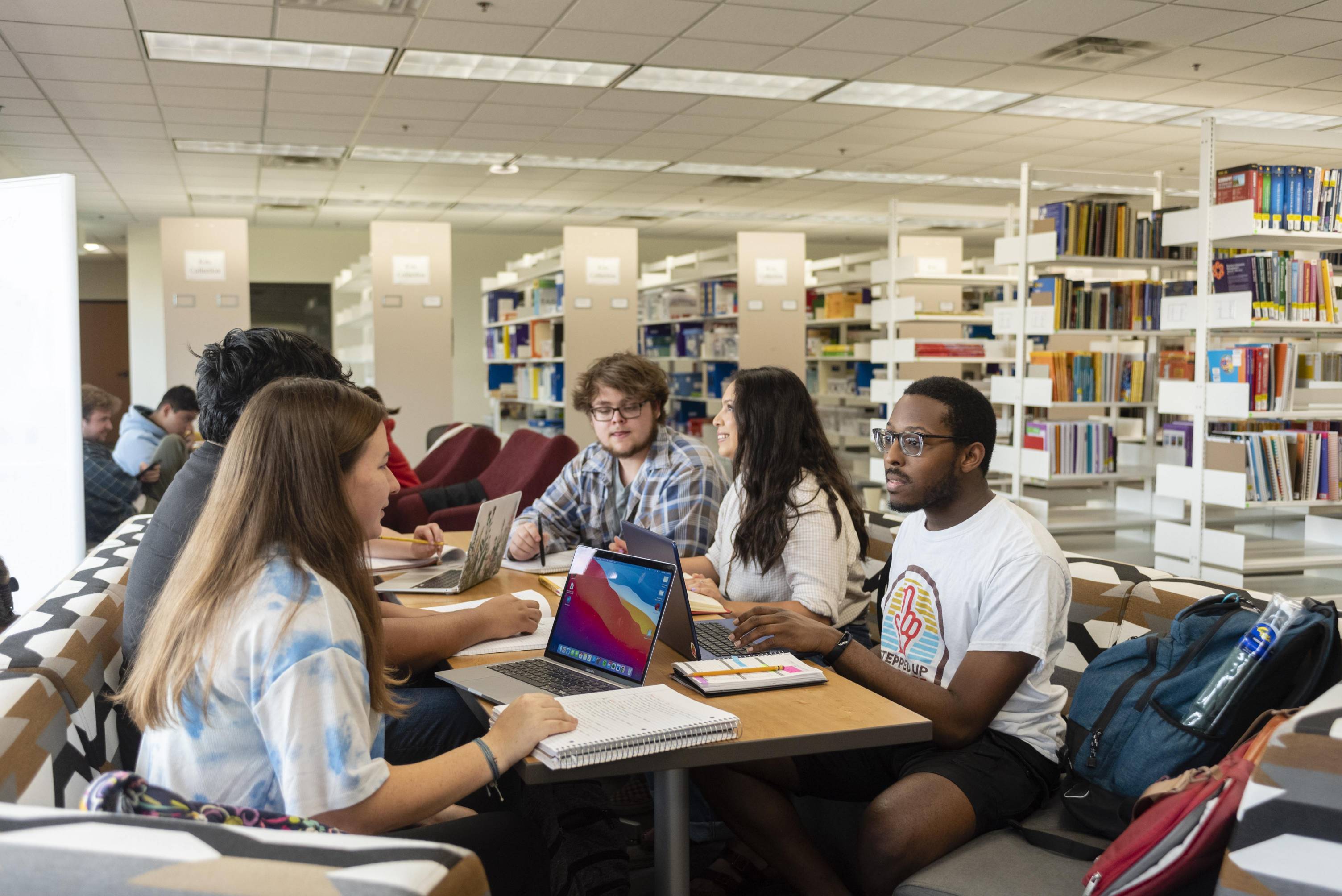 A group of diverse college students is gathered around a table in a bright library, engaged in studying and discussion. Several students have open laptops and notebooks. The background features rows of bookshelves filled with books.