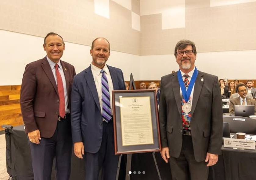 Three professionals in suits stand in a formal setting. The professional on the left wears a burgundy suit with a red tie and smiles at the camera. The professor in the center wears a navy suit with a striped tie, holding a framed document and smiling. The professor on the right wears a dark suit with a medal around their neck and glasses, smiling at the camera. Other people are visible in the background, seated at tables.