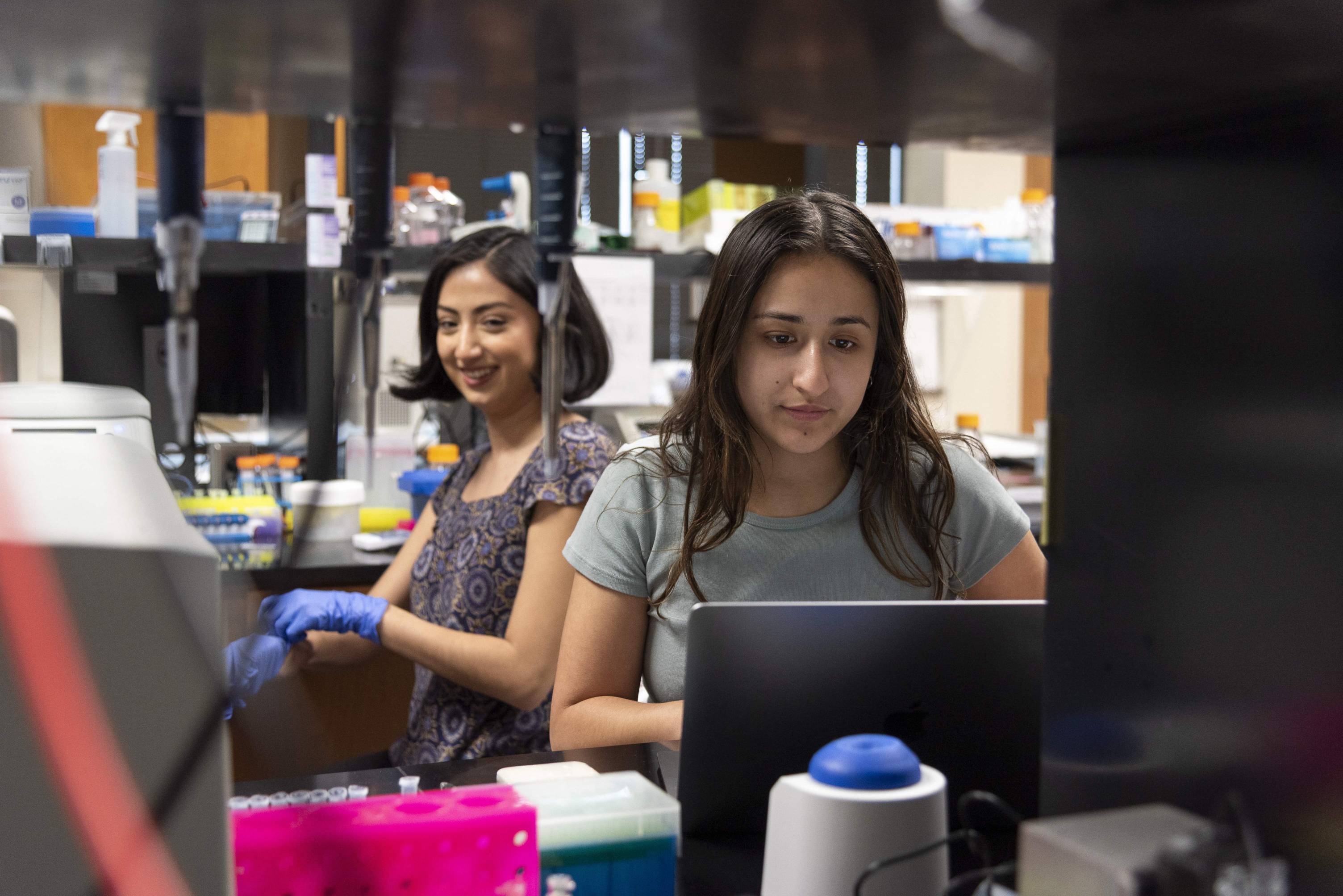 Two students are in a lab setting. In the foreground, a student with dark hair in a light green t-shirt is focused on a laptop. In the background, another student with a shorter haircut and blue gloves is visible, smiling and working at a lab bench with various scientific equipment.