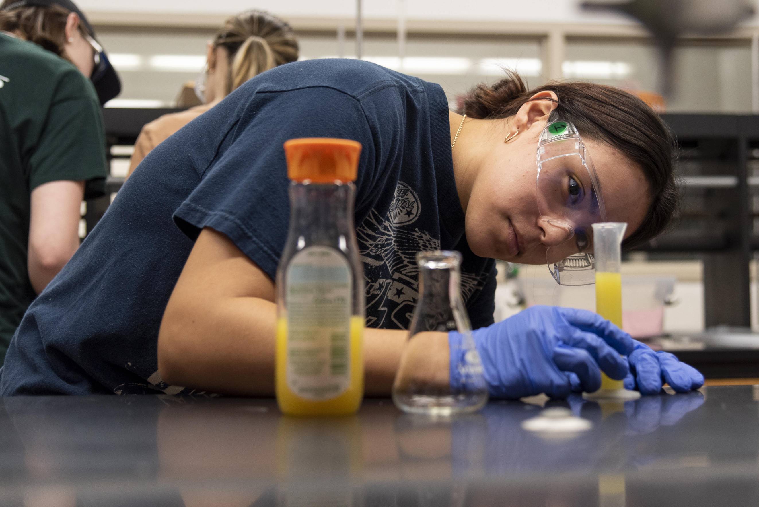  A student wearing safety goggles and blue gloves leans over a lab bench, intently observing a yellow liquid in a graduated cylinder. A bottle of orange juice and an empty beaker are also on the bench. Other students are blurred in the background.