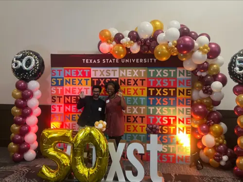 Two people in business clothing pose in front of a colorful backdrop, surrounded by balloons.