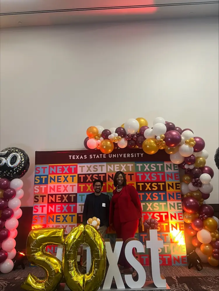 Two people in business clothing pose in front of colorful backdrop, surrounded by balloons.