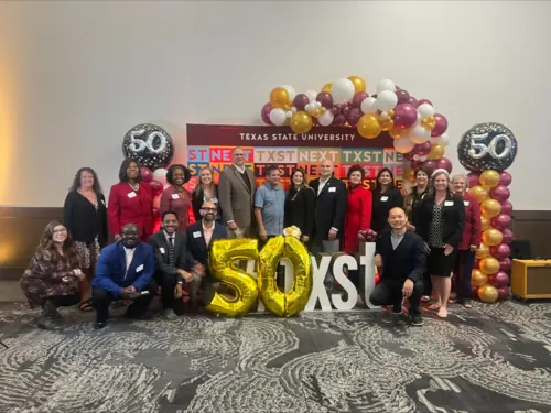 A group of people in business clothing standing in front of a maroon and gold backdrop, covered in balloons that say "50."