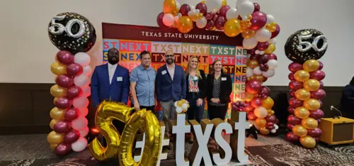 Several people pose in front of a colorful backdrop, surrounded by balloons. 