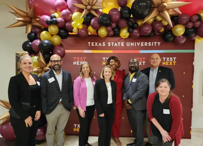 Several people pose in front of a maroon backdrop, balloons overhead. 