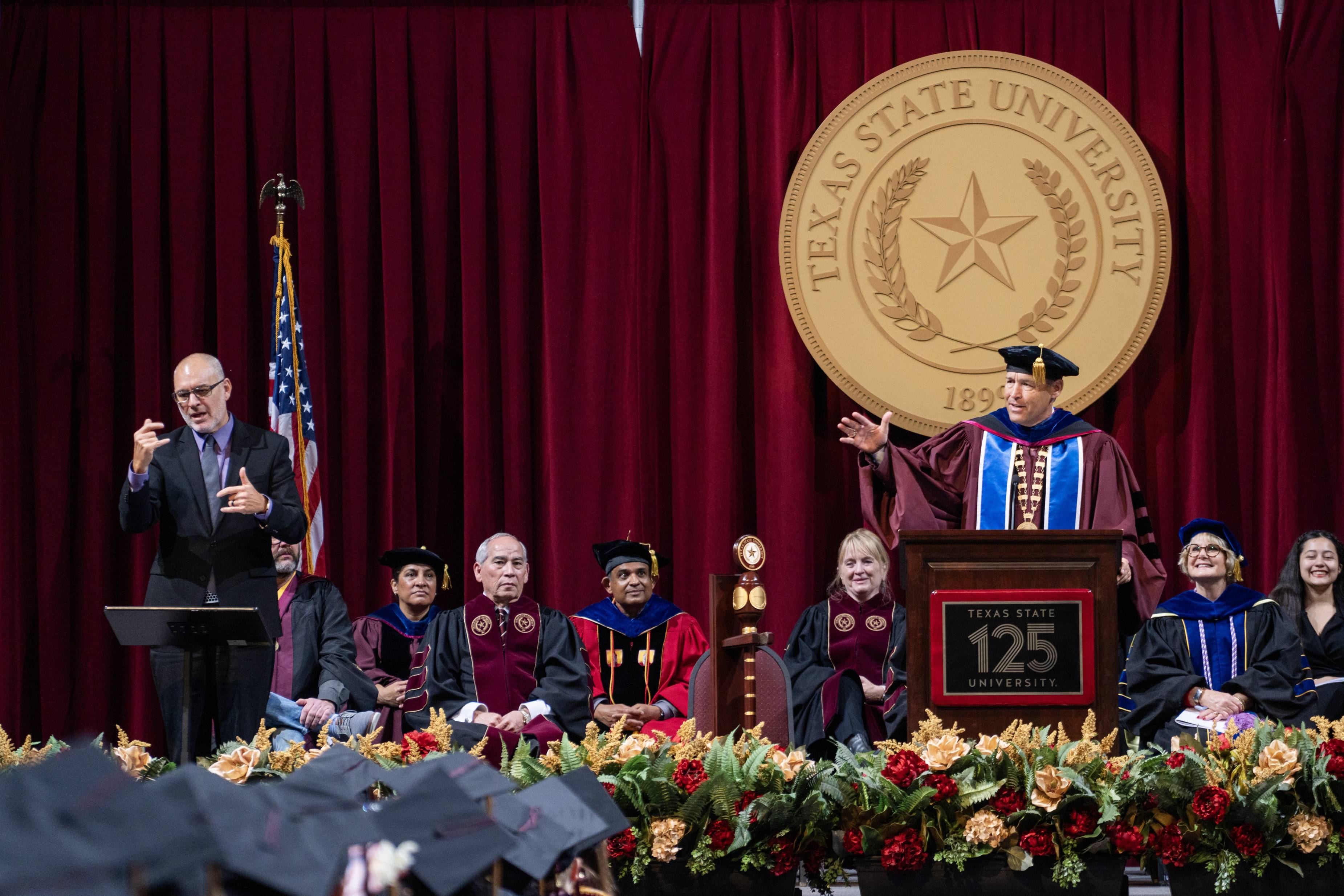 President Damphousse stands at a podium delivering a commencement speech. An interpreter is at a separate podium providing interpreting services.