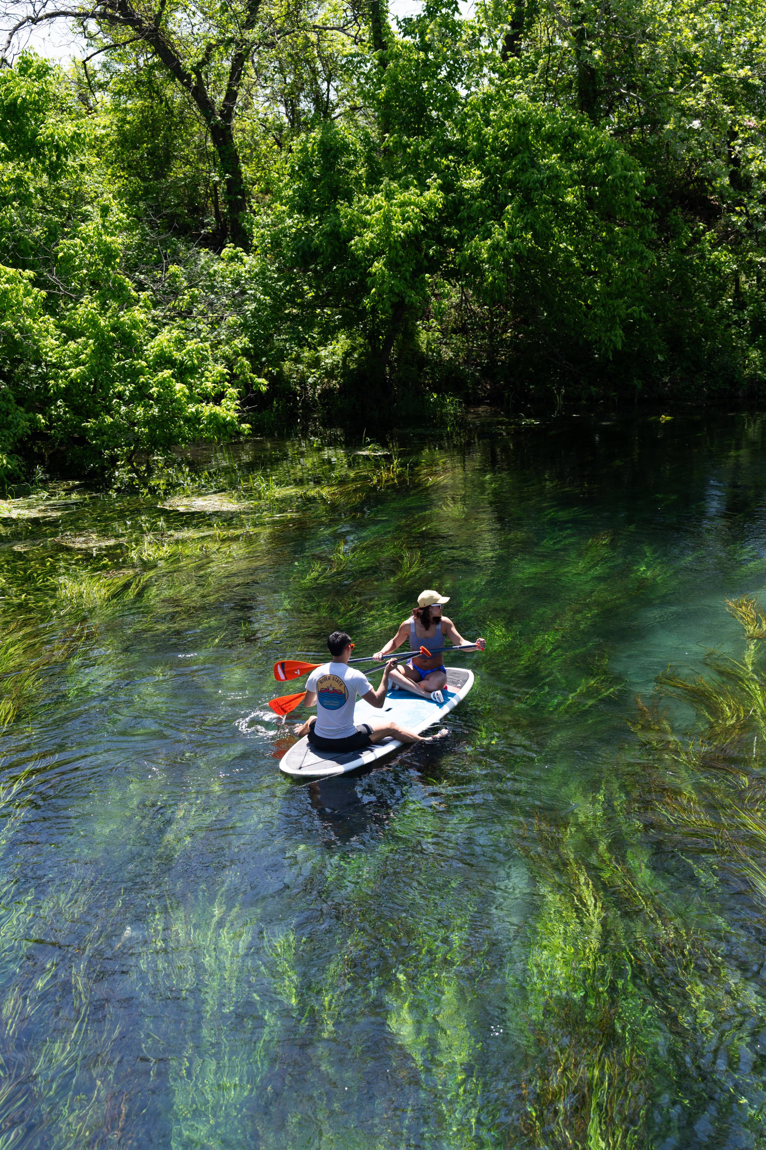 Two students navigate the San Marcos River on a paddle board. 